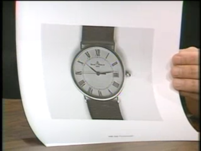 A hand holding up a piece of paper with the printed gray-scale image of a wrist watch.