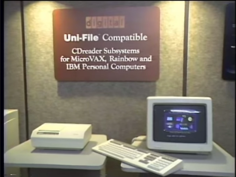 A trade show display of the DEC CDreader for MicroVAX, Rainbow, and IBM Personal Computers. The CD-ROM drive is a larger rectangular unit connected to a computer with a separate keyboard and monitor.