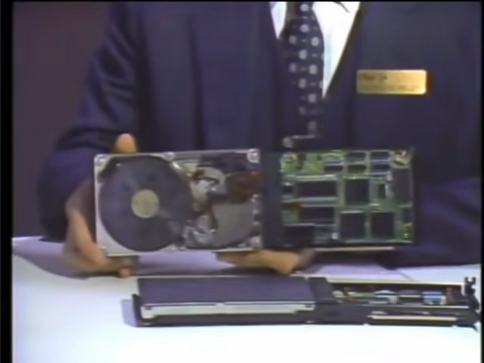 A man in a suit holding a hard disk drive mounted to a circuit board. The unit is designed to be inserted into an expansion slot on an IBM PC or compatible machine.