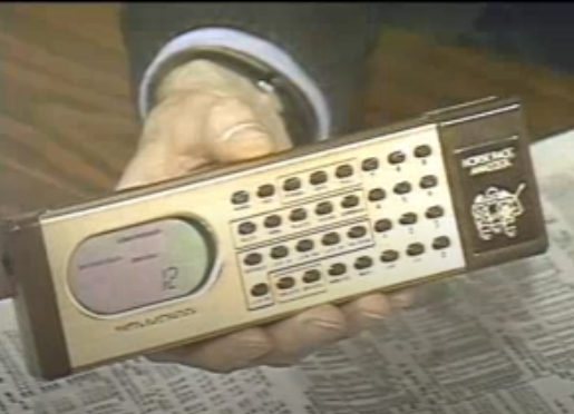 Stewart Cheifet holding the Mattel Electronics Horse Race Analyzer, a long rectangular-shaped calculator with an LCD screen on the left side, on the set of “Computer Chronicles” in 1987.
