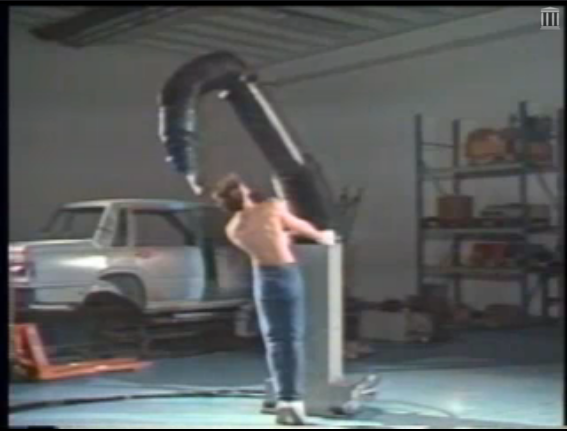 A shirtless man dancing with a giant robotic arm in an auto garage.