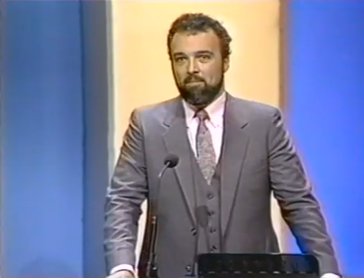Paul Schindler, wearing a gray three-piece suit, standing behind a contestant’s podium on the set of “Jeopardy!