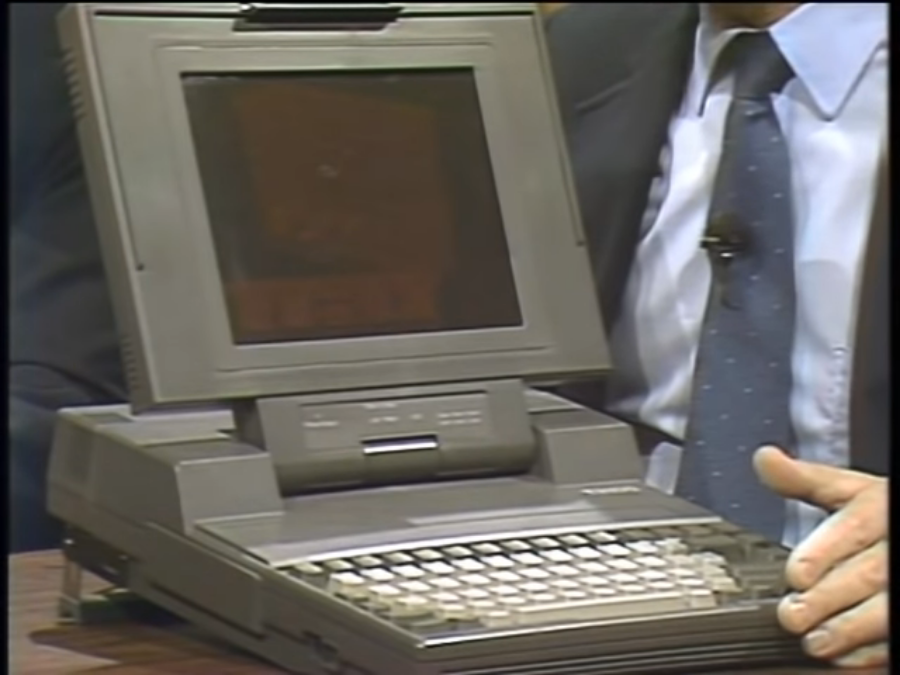 The Toshiba T3100 computer on the set of “Computer Chronicles.” The machine has a gray color. The orange plasma display screen is open, revealing a keyboard underneath. The laptop is perched at a slight angle.