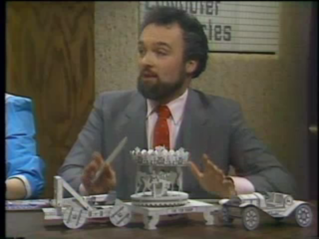 Paul Schindler sitting at the desk on the set of “Computer Chronicles.” On the desk in front of him are three paper models of a catapult, carousel, and open-air car.