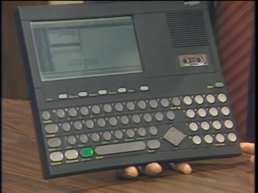 A close-up shot of Stewart Cheifet holding a Convergent Technologies Workslate portable computer. The Workslate was a large, flat black tablet with a small LCD screen built-in to the top left corner, a micro-cassette drive built-in to the top right corner, and a keyboard composed of small circular keys covering the lower half.